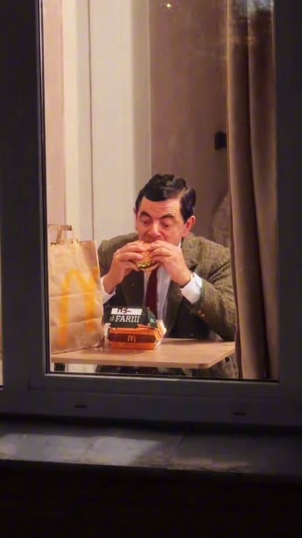 A person eats a burger behind a warmly lit apartment window at night.