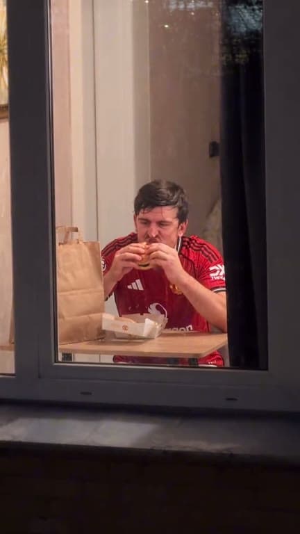 A person enjoys a burger at a table inside a lit window after dark.