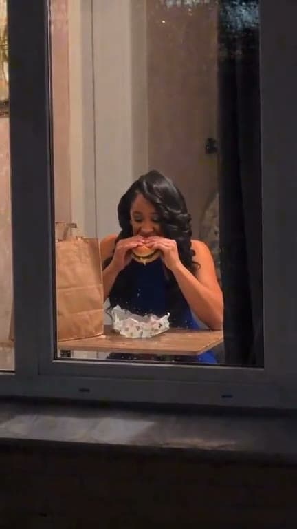 A person takes a late-night burger bite behind a lit window.